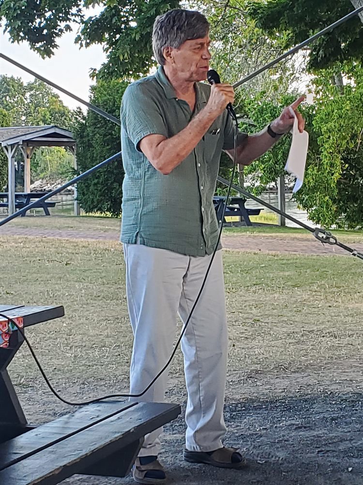 Peter Somssich speaking into a hand-held microphone and holding his notes on the lawn at Four Tree Island